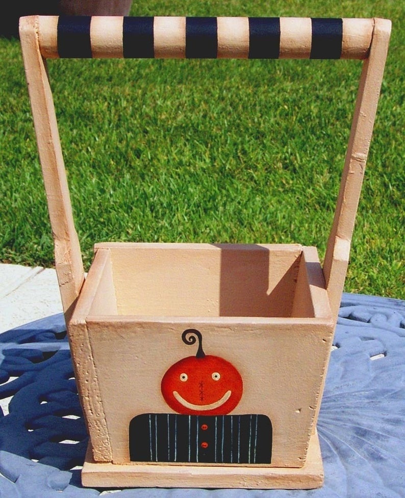 May include: A wooden pumpkin-themed tote box with a black and white striped handle. The box is painted with a smiling pumpkin face and a black and white striped shirt.