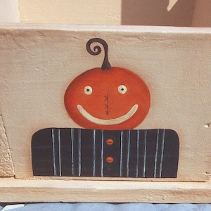 May include: A wooden box with a painted pumpkin face. The pumpkin is orange with a black stem and a stitched smile. The box is painted a light brown color.