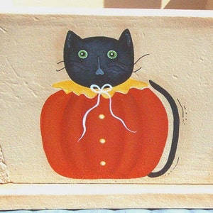 May include: A hand-painted wooden planter box with a black cat wearing a pumpkin costume. The cat has green eyes and a white bow tie. The planter box is painted in a light beige color.