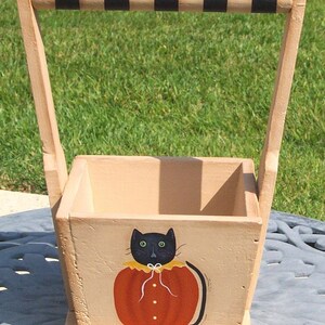 May include: A wooden tote with a black and white striped handle. The tote is painted with a black cat wearing an orange pumpkin costume.