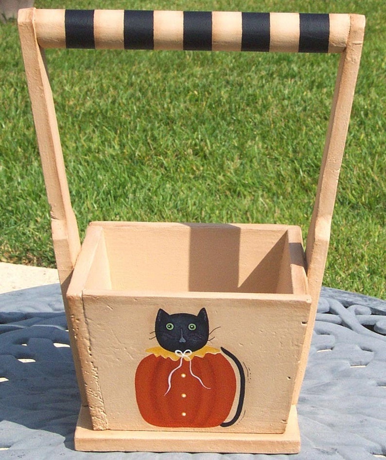 May include: A wooden tote with a black and white striped handle. The tote is painted with a black cat wearing an orange pumpkin costume.