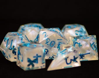 May include: A collection of clear, iridescent dice with blue numbering and decorative symbols. The dice are various polyhedral shapes, including six-sided and twenty-sided dice, suitable for tabletop gaming.