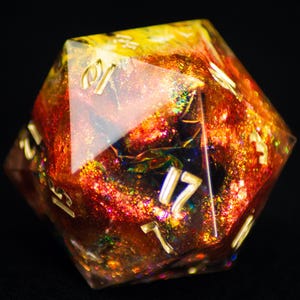 May include: A multi-faceted, resin D20 die with a fiery, galaxy-like interior. The die features gold numbering and a mix of orange, yellow, and red hues with iridescent glitter. The overall effect is a vibrant, eye-catching design.
