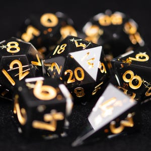Puede incluir: Un conjunto de dados poliédricos en negro con números de color dorado y escamas de oro incrustadas. La colección incluye varias formas como un dado de veinte caras, un cubo y un dado de cuatro caras, todos con bordes afilados y números claros.