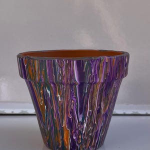 May include: A small terracotta plant pot with an abstract paint design. The pot is decorated with vertical streaks of purple, orange, white, and green paint. The inside of the pot is a terracotta colour.