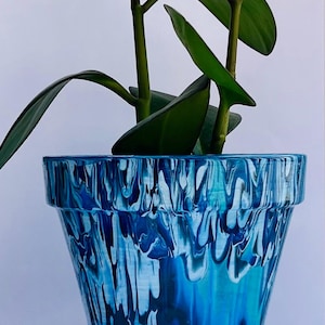 May include: A blue and white swirled ceramic plant pot with a matching saucer. The pot contains a green leafy plant. The pot has a glossy finish and a unique, artistic design. The plant pot is a decorative item for home decor.
