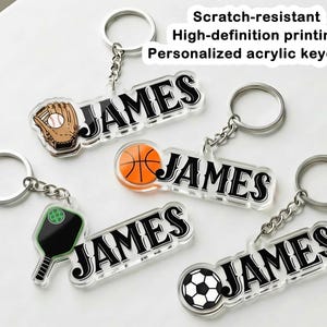 Könnte beinhalten: Transparente Acryl-Schlüsselanhänger mit dem Namen "JAMES" in Schwarz und Weiß, jeweils mit einem anderen Sportmotiv: Baseballhandschuh, Basketball, Pickleballschläger und Fußball. Die Schlüsselanhänger haben silberne Schlüsselringe und Ketten. Der Text auf dem Bild lautet "Scratch-resistant High-definition printing Personalized acrylic keychain".