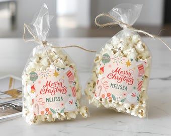 May include: Two clear cellophane bags filled with popcorn, tied with twine. Each bag has a label with "Merry Christmas" in red script, the name "Melissa", and festive illustrations of ornaments and a snowman.