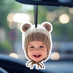 May include: A car air freshener featuring a smiling baby wearing a brown and white knit hat with bear ears. The air freshener is hanging from a black string, with a blurred background.