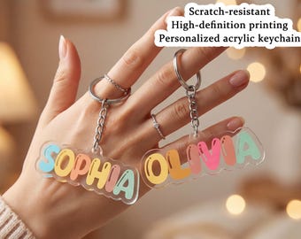 Może przedstawiać: Dwa spersonalizowane breloki akrylowe z imionami "SOPHIA" i "OLIVIA" w kolorowych literach. Posiadają srebrne metalowe kółka i łańcuszki. Widoczny jest tekst "Scratch-resistant High-definition printing Personalized acrylic keychain".