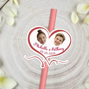 May include: A pink straw with a heart-shaped sticker featuring two smiling faces and the names "Michelle & Anthony" with the date "05.29.2025" in red script. White flower petals surround the straw.