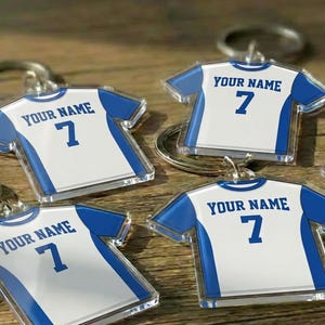 May include: Keyrings with a white and blue jersey design, displaying the text "YOUR NAME" and the number "7". The keyrings are made of clear acrylic and have a silver keyring attached. The jerseys are on a wooden surface.