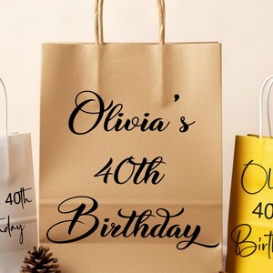 May include: Three gift bags in beige, white, and yellow. The beige bag has the text "Olivia's 40th Birthday" in black script. The white and yellow bags also have the same text. The bags have handles.
