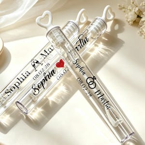 May include: Three clear plastic bubble wands with white heart-shaped handles. Each tube features black printed text reading Sophia and Martin, the date 08.02.28, and icons including champagne flutes, a red heart, and interlocking wedding rings.