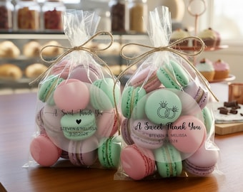 May include: Two clear cellophane bags tied with twine, filled with pastel macarons. The macarons are pink, green, and lavender. Each bag has a circular label with the text "A Sweet Thank You" and a wedding date.