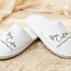 May include: Two white, plush slippers with the text "Your Custom Logo/Image/Text" printed in black script. The slippers are on a beige carpet, with a soft, cream-coloured fabric in the background.