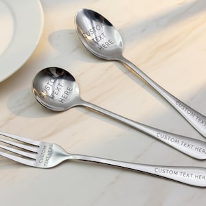 May include: A set of three stainless steel utensils: a spoon, a smaller spoon, and a fork. Each utensil has the text "CUSTOM TEXT HERE" engraved on the handle and the spoon bowls. The utensils are on a light-colored surface.