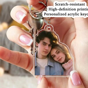 Op de afbeelding: Een heldere acryl sleutelhanger met een foto van een stel. De sleutelhanger is bevestigd aan een zilveren sleutelring met sleutels. Tekst op de sleutelhanger luidt: "Krasbestendig, High-definition printing, Gepersonaliseerde acryl sleutelhanger."