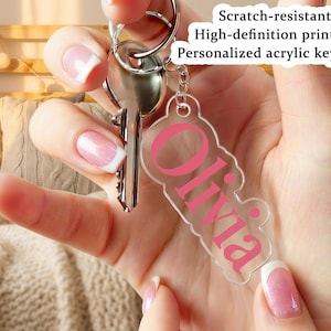 Puede incluir: Un llavero de acrílico transparente con el nombre "Olivia" en letras rosas. El llavero está unido a un llavero plateado con llaves. El texto de la imagen dice "Resistente a los arañazos, Impresión de alta definición, Llavero de acrílico personalizado."