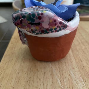 May include: A small terracotta-colored clay pot featuring a hand-painted snake with a multi-colored spotted pattern wrapped around the rim. A blue fabric flower is tucked inside the pot, extending upwards from the center of the snake's coil.