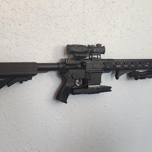 May include: A black rifle with a scope and a torch attached to the bottom. The rifle is mounted on a white wall.