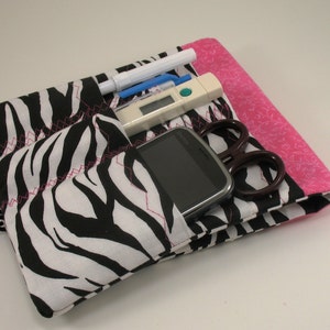May include: A black and white zebra print fabric pouch with pink trim. The pouch has multiple pockets and is filled with various items, including a cell phone, a thermometer, a pen, and a pair of scissors.