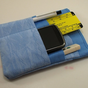 May include: A light blue fabric pouch with a zippered pocket and a flap closure. The pouch contains a smartphone, a pen, a ruler, and a small white tool.
