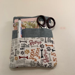 May include: A grey and white fabric pouch with a dog bone and paw print pattern. The pouch has multiple pockets and is filled with a pen and a pair of scissors. The text on the pouch includes "woof", "bow", "ruff", "sniff", "groom", and "grrr".