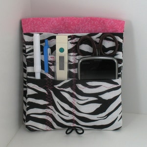 May include: A black and white zebra print fabric organizer with pink stitching. The organizer has multiple pockets and is filled with various items, including a white thermometer, a pair of scissors, a blue pen, a white pen, and a black cell phone.