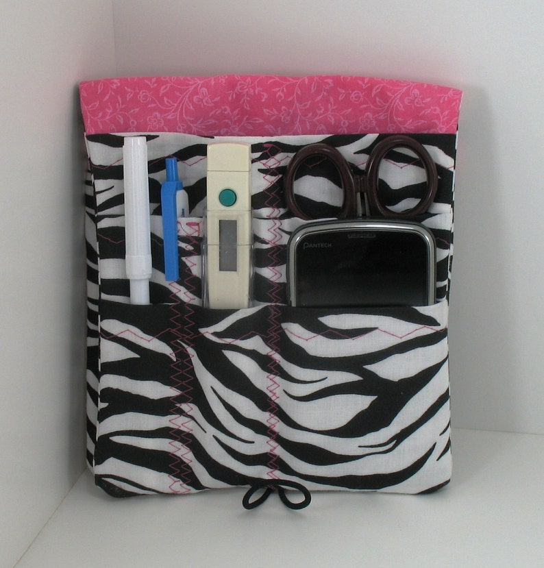 May include: A black and white zebra print fabric organizer with pink stitching. The organizer has multiple pockets and is filled with various items, including a white thermometer, a pair of scissors, a blue pen, a white pen, and a black cell phone.