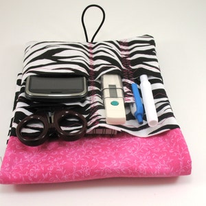May include: A black and white zebra print fabric pouch with a pink floral fabric lining. The pouch has multiple pockets and is designed to hold items like a phone, thermometer, scissors, and pens.