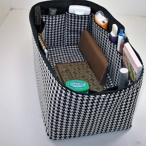 Pode incluir: Organizador de tecido com padrão preto e branco com vários bolsos para uma bolsa de mão. O organizador está cheio de vários itens, incluindo uma carteira, canetas e um pequeno caderno.