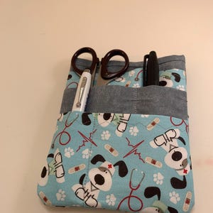 May include: A blue fabric medical tool organizer with a pattern of cartoon dogs, bandages, and stethoscopes. The organizer holds scissors, a pen, and a silver pen. The organizer has a gray pocket.