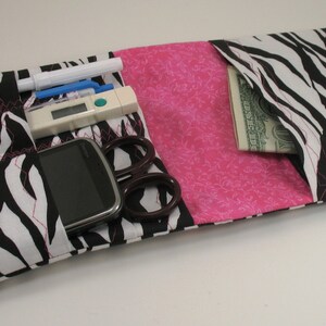 May include: A black and white zebra print fabric pouch with a pink fabric lining. The pouch has multiple pockets and is open to reveal a cell phone, a thermometer, a pen, a pair of scissors, and a dollar bill.