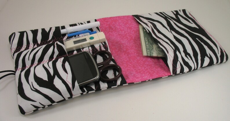 May include: A black and white zebra print fabric pouch with a pink fabric lining. The pouch has multiple pockets and is open to reveal a cell phone, a thermometer, a pen, a pair of scissors, and a dollar bill.