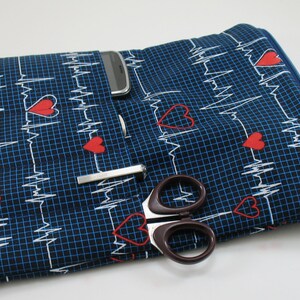 May include: A blue fabric organizer with a zipper closure and multiple pockets. The fabric has a pattern of white EKG lines and red hearts. The organizer has a stethoscope, scissors, a pen, and a cell phone in the pockets.