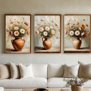 May include: Set of three framed floral paintings. Each features a terracotta vase filled with a bouquet of white, pink, and orange flowers. The artwork has a soft, painterly style with a neutral background and wooden frames, suitable for home decor.