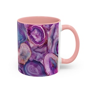 May include: A white ceramic mug with a pink interior and handle. The exterior features a vibrant, abstract design of purple and blue agate stone patterns. The mug is designed for hot or cold beverages.