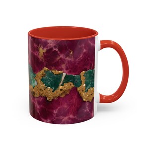 May include: A white ceramic mug with a red interior and handle. The exterior features a marbled design in shades of burgundy and gold, with a band of green and gold across the center. The mug is designed for hot beverages.