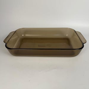 May include: A rectangular baking dish made of translucent brown glass. The dish features integrated handles on both short ends and a smooth, polished rim. Embossed text on the interior base reads "baker" along with other faint markings.