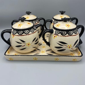 以下が含まれることがあります： 黒い取っ手と蓋が付いた、黒と黄色の花柄が施された、クリーム色のセラミックティーカップ4客のセット。 カップは、同様に花の模様で飾られた、お揃いの長方形のトレイに置かれています。 紅茶を出すためのセットです。