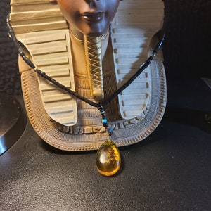 May include: A black cord necklace with a teardrop-shaped amber pendant. The amber pendant is a translucent golden-yellow color with an insect inside. The necklace has small black and gold beads and is displayed in front of a bust of an Egyptian pharaoh.