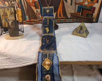 May include: A unique, handmade necklace with a denim pendant. The pendant features gold-colored embellishments, including a star, ankh, and a geometric design with stones. The necklace is displayed on a black mannequin head against a backdrop of Egyptian-themed artwork.