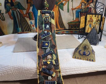 May include: A handmade pendant necklace with a long, rectangular pendant. The pendant features a camouflage pattern with denim and stone accents, and a gold-colored trim. The necklace is displayed on a brown bust, with an Egyptian-themed tapestry in the background.