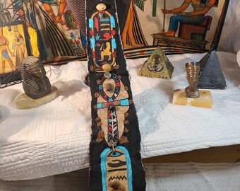 May include: A black, rectangular pendant with Egyptian hieroglyphs in red, blue, and gold, suspended from a silver chain. The pendant features an ankh symbol and other ancient Egyptian motifs. The background includes Egyptian-themed tapestries and decorative items.