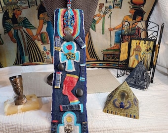 May include: A handcrafted denim necklace with Egyptian-themed embellishments. The pendant features ankh symbols, hieroglyphs, and other decorative elements in various colors. The necklace is displayed on a mannequin bust.