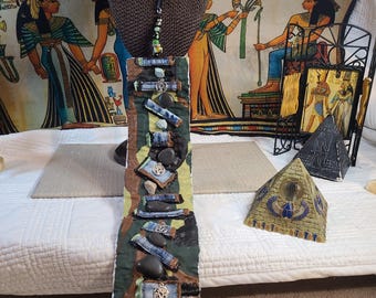 May include: Handmade necklace with a long, rectangular pendant. The pendant features a camouflage pattern, denim accents, and small stones. Silver-toned charms with a tree design are also present. The necklace is displayed on a brown bust.