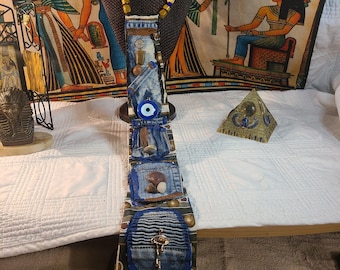May include: A unique, handmade necktie with a denim patchwork design, adorned with beads, a key pendant, and a silver-toned oval embellishment. The tie is displayed on a mannequin head, with an Egyptian-themed backdrop.