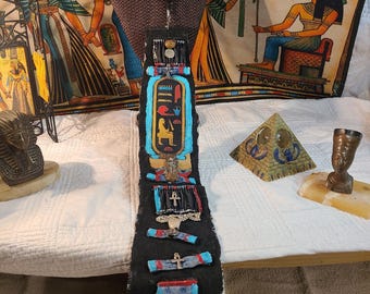 May include: A black, handcrafted necktie with Egyptian hieroglyphic designs. The tie features a central panel with a turquoise border and gold hieroglyphs. Additional embellishments include small metal charms and colorful accents.