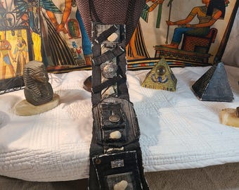 May include: A unique, handmade necklace featuring a dark denim pendant adorned with stones and embellishments. The pendant is attached to a black beaded chain and includes a striped tie at the bottom. The necklace is displayed on a mannequin.
